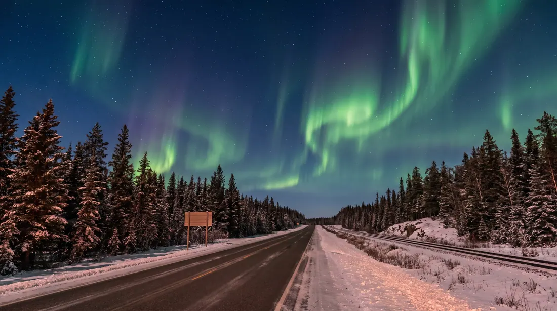 A northern highway and a rail line through spruce forest under green aurora