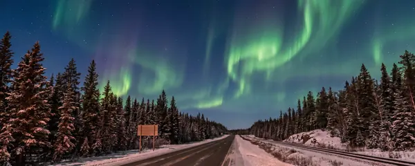 A northern highway and a rail line through spruce forest under green aurora