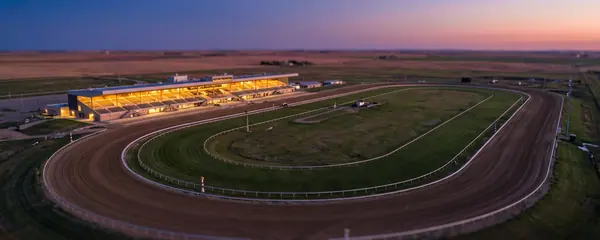 An empty dirt racetrack and a lit grandstand at dusk, seen from above