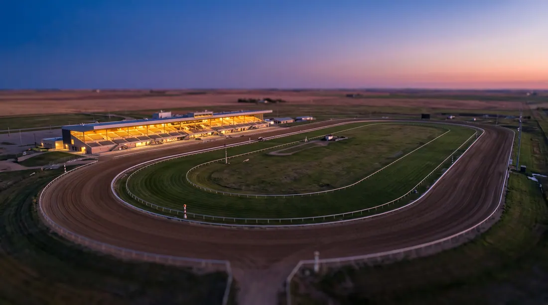 An empty dirt racetrack and a lit grandstand at dusk, seen from above